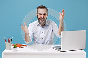 Angry shocked young bearded man in light shirt sit work at desk with pc laptop isolated on blue background. Achievement