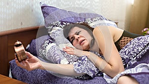 Angry sleepless woman in bed with an alarm clock in her hands, the concept of an unloved job