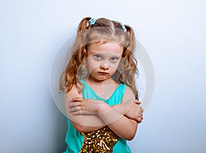 Angry small girl kid with closed gestured folded arms looking serious on blue background. Closeup