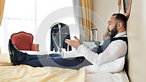 Angry stylish male businessman lying on bed at vintage hotel room having mobile conference