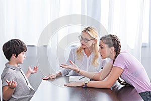 angry teenage girl pointing by hand on little brother doing shrug gesture on family therapy session by female