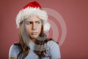 Angry unhappy caucasian girl teenager standing in santa claus red hat looking aside over pink background. Studio portrait. Anti