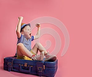 Angry upset little kid sitting on the suitcase having upset facial expression wants to travel. Child in hat with hands