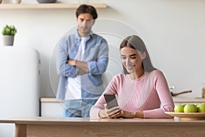 Angry upset offended young husband looking at wife with smartphone at kitchen interior, empty space