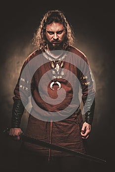 Angry viking with sword in a traditional warrior clothes, posing on a dark background.