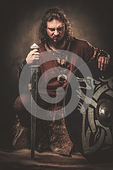 Angry viking with sword in a traditional warrior clothes, posing on a dark background.