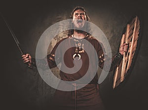 Angry viking with sword in a traditional warrior clothes, posing on a dark background.