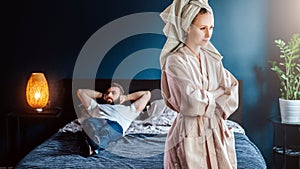 Angry woman in bathrobe stands in bedroom,her arms crossed over chest. Man is lying on bed .Couple having problems.