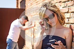 Angry woman looking phone and man posing on wall