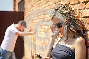 Angry woman looking phone and man posing on wall