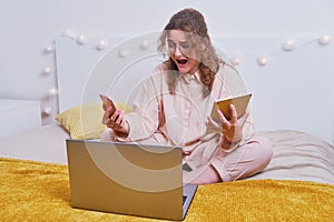 Angry woman screaming with a phone and a tablet in her hands on a home bed. Problems working online during isolation due to the