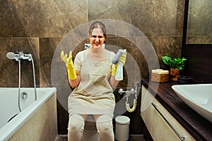 Angry woman sits on the toilet while cleaning the bathroom. Disinfection with yellow rubber gloves in the washroom