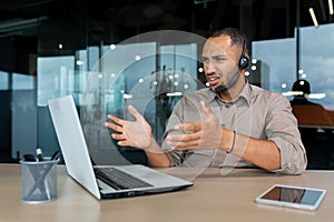 Angry worker with video call headset talking to customer, hispanic man explaining angry and upset, businessman inside