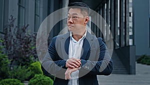 Angry worried middle-aged man 40s Asian Korean businessman standing in city looking at wristwatch check time waiting