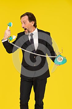 Angry, yelling middle aged business man in to old fashion telephone handset holding it in hands isolated on yellow