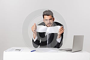 Angry young bearded man work at desk with laptop isolated over white background. Screaming tearing paper documents