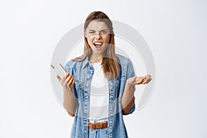 Angry young blond woman staring at camera annoyed, holding smartphone, complaining at bothering news online, standing