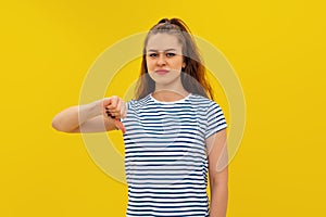 Angry young brunette woman in striped t shirt, looking upset, showing thumb down. Indoor studio shot on yellow background