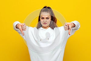 Angry young brunette woman in white casual style sweatshirt, looking upset, showing thumbs down. Indoor studio shot on yellow