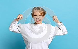 Angry young caucasian girl wearing white pullover clenches fists isolated on blue background. Concept of emotions