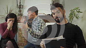 Angry young Caucasian man yelling at his wife at the background, woman crying. Professional male psychologist taking off