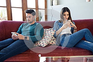 Angry young couple ignoring each other using phone after an argument while sitting on sofa at home