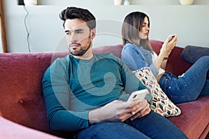Angry young couple ignoring each other using phone after an argument while sitting on sofa at home