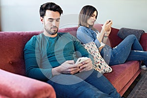 Angry young couple ignoring each other using phone after an argument while sitting on sofa at home