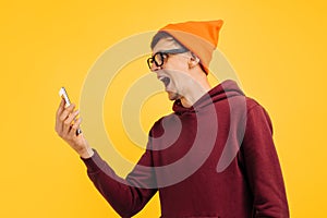Angry young guy in an orange hat with glasses, red sweater shouts angry into phone, talking on phone in a loud voice