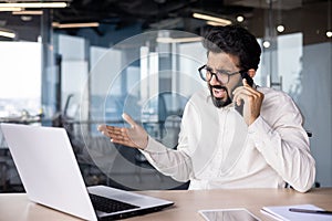 Angry young Indian male businessman sitting in the office at the desk and emotionally talking on the phone