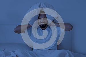 Angry Young Indian Man Covering Ears With Pillow While Sitting In Bed