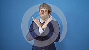 Angry young man in business suit expresses rejection with firm crossed arms gesture and negative sign, isolated on blue background