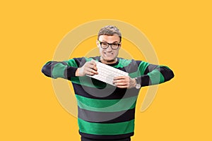 Angry young man holding a keyboard in his hands and trying to break it on yellow background. Studio photo