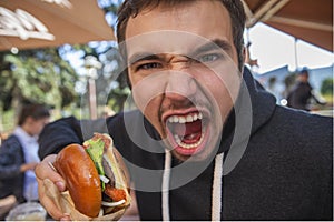 Angry young man looking at the camera with his mouth wide open. He is holding his hamburger at his right hand. Bad taste concept