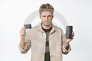 Angry young man shows credit card and mobile phone screen, not enough fonds on bank account, showing mobile banking app