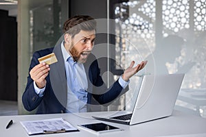 Angry young man in a suit working in the office, sitting at a desk, holding a credit card, looking worriedly at the