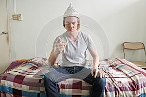 Angry young man with tin foil hat pointing at camera in the bedroom