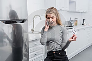 Angry young woman holding card with lettering home inspection and yelling on smartphone