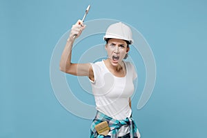 Angry young woman in protective helmet hardhat hold adjustable wrench isolated on blue wall background. Instruments