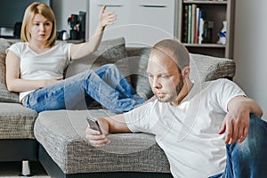 Angry young woman sitting at the sofa talking to her husband while he is using his cellphone in their living room.