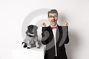 Animals, party and celebration concept. Happy young man in suit and puppy in pet cosume standing over white background