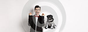Animals, party and celebration concept. Happy young man in suit and puppy in pet cosume standing over white background