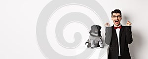 Animals, party and celebration concept. Happy young man in suit and puppy in pet cosume standing over white background