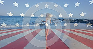 Animation of flag over america over happy caucasian woman walking towards sea at beach