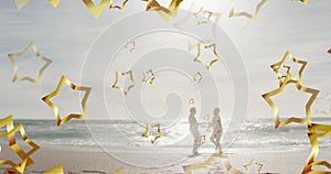 Animation of gold christmas stars over happy silhouetted couple dancing on sunny beach