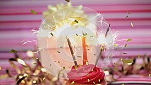 Animation of gold confetti over hand holding match lighting candle and sparklers on birthday cupcake