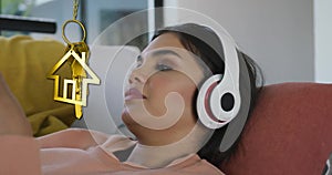 Animation of gold house key and key fob over biracial woman using headphones at home