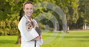 Animation of happy caucasian female vet holding yorkshire terrier pet dog, over trees in park