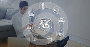 Animation of icons in hexagon over asian man sitting on ground with laptop and using cellphone