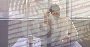 Animation of plants and rain on window over tired caucasian businessman using laptop at office desk